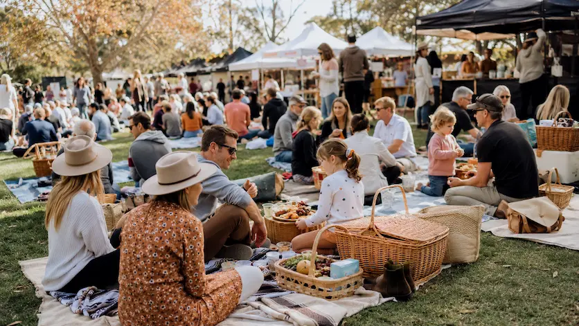 Families and visitors enjoying a big picnic at a GourMay-style food festival in Imbil, Queensland, with picnic rugs, baskets and food stalls.